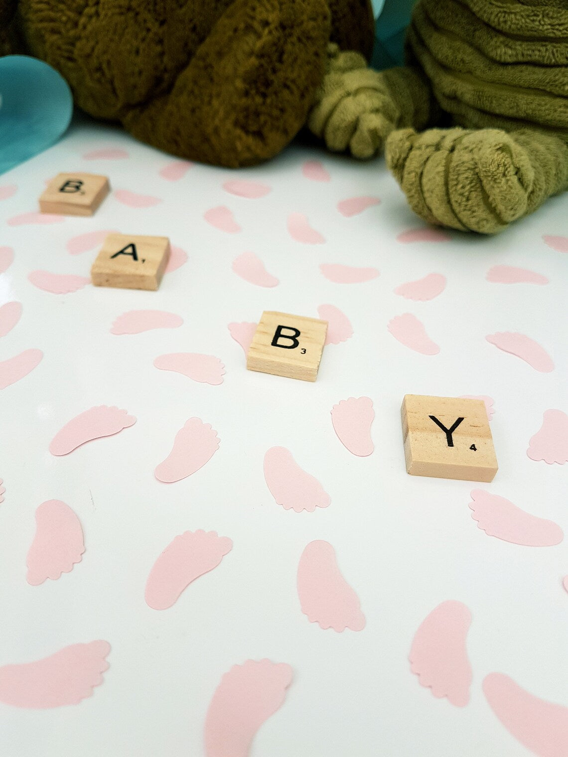Pastel pink baby feet shaped confetti pieces are scattered on a white background with wooden scrabble tiles reading BABY diagonally across the photo. Cuddly toys are seen in the background