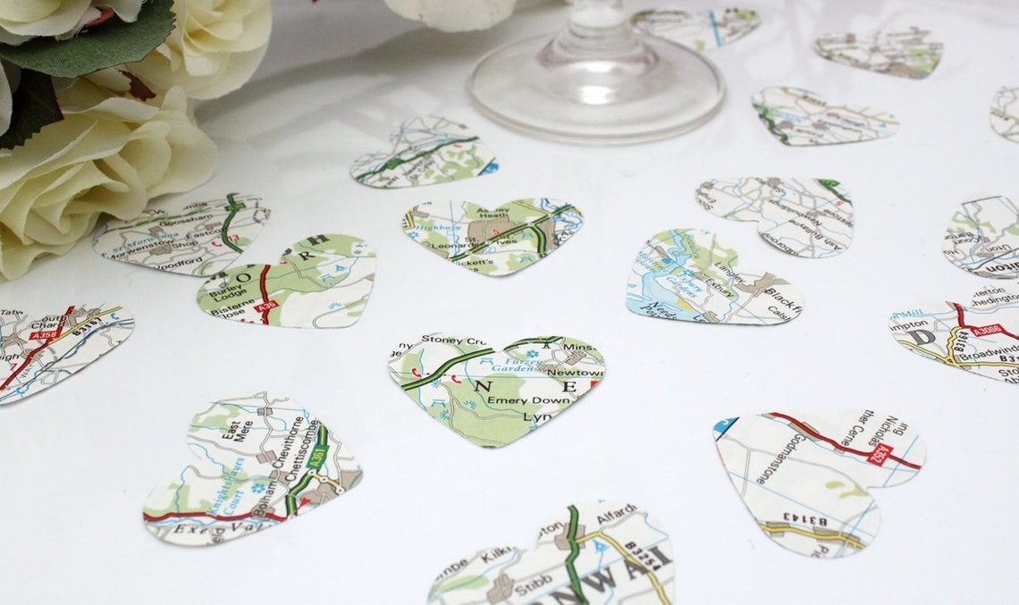 Map paper confetti pieces in heart shapes are scattered on a white background with a champagne flute and purple orchid bouquet in the background. The paper confetti is primarily white with green, blue and red colours