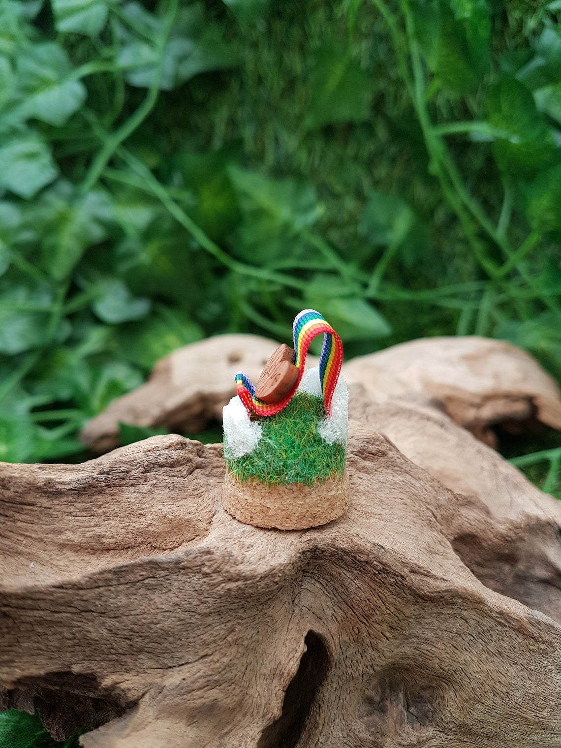 Miniature glass bell jar base with a curl of rainbow ribbon with miniature clouds at the start and end of the ribbon at the back and front. A small wooden heart with sits on the ribbon, engraved with the word LOVE
