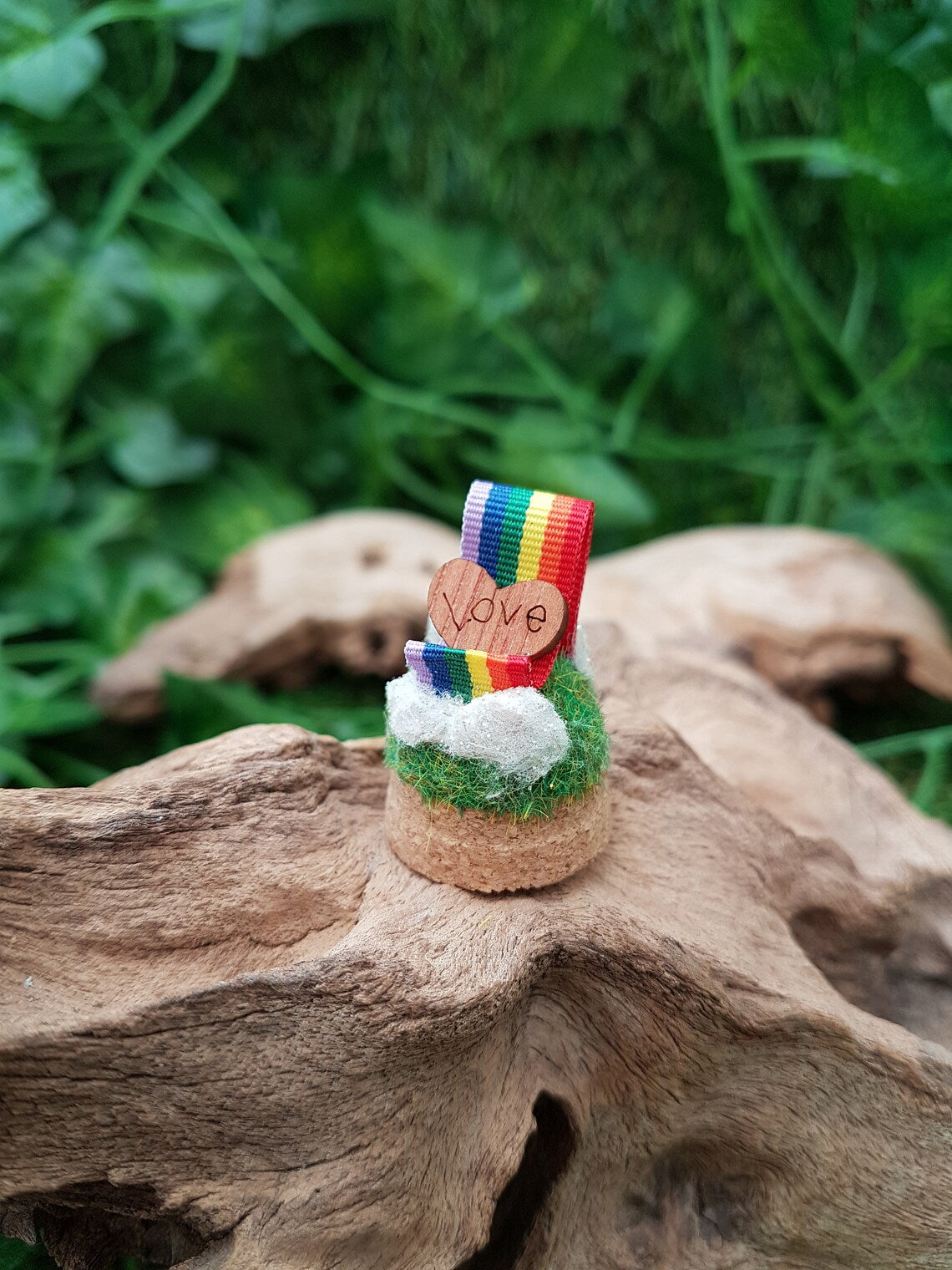 Miniature glass bell jar base with a curl of rainbow ribbon with miniature clouds at the start and end of the ribbon at the back and front. A small wooden heart with sits on the ribbon, engraved with the word LOVE