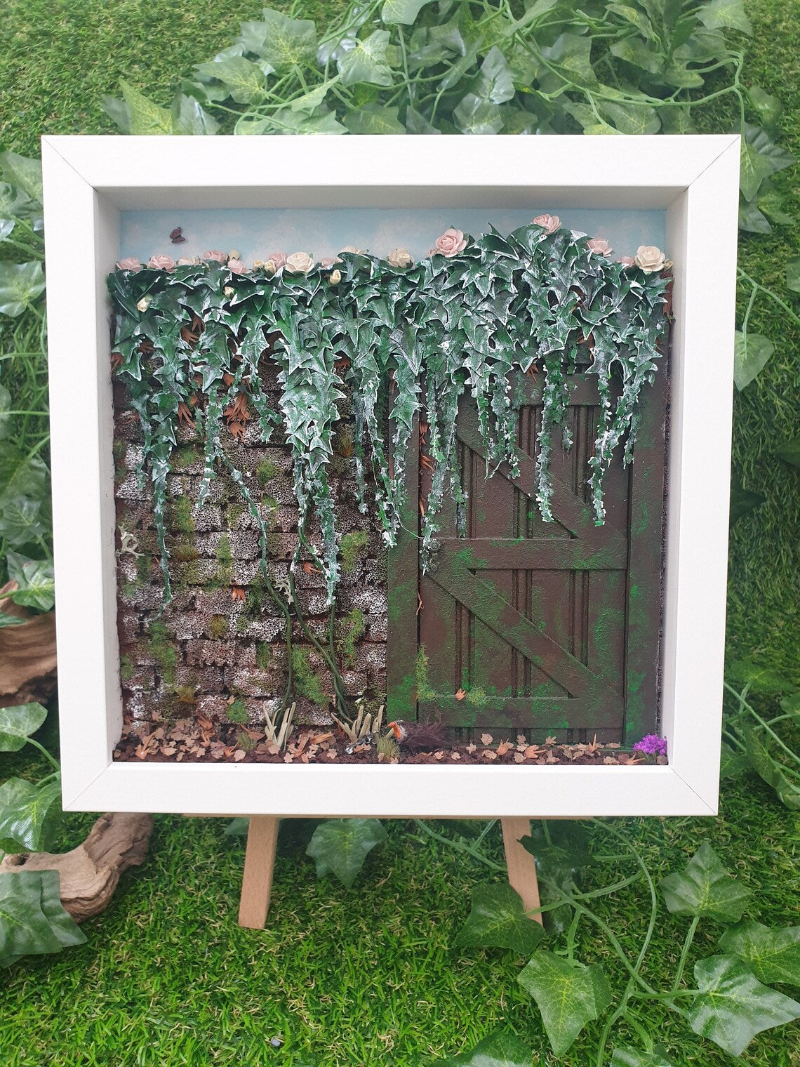 A square white box frame on an easel contains a miniature 1:12 diorama consisting of a weathered door in a brick wall with ivy hanging down over the wall and door. There is a tiny robin satin front of the door