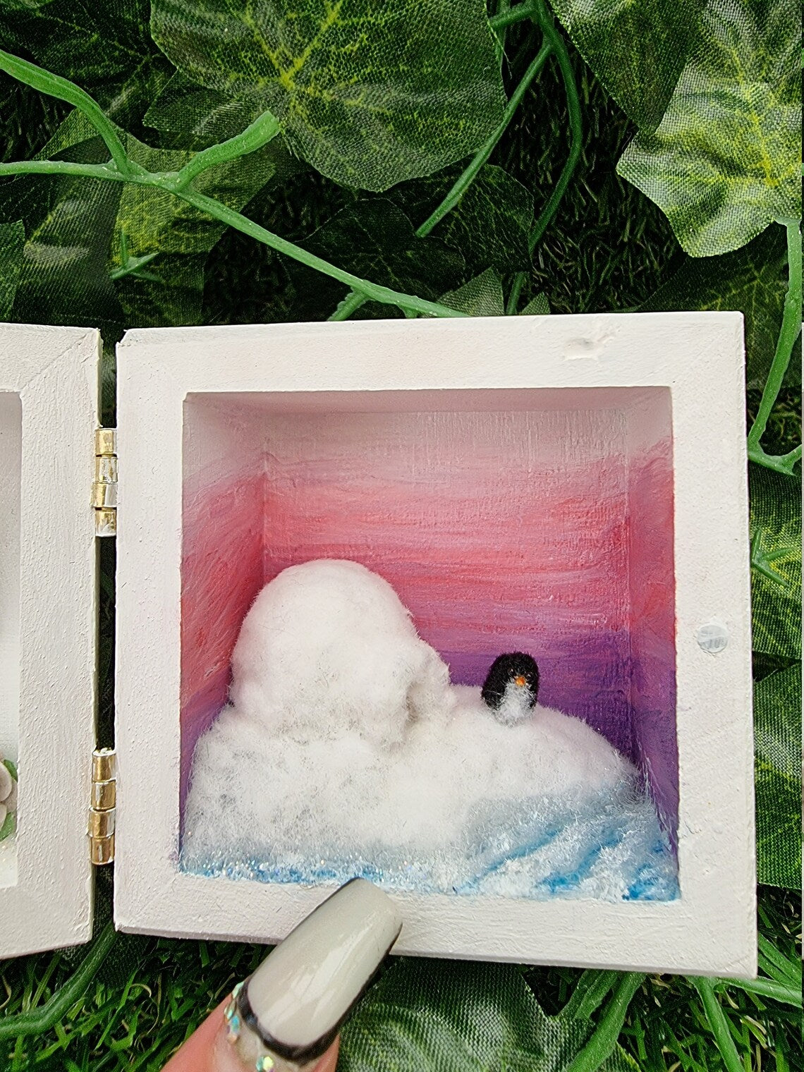 A small white square box, open like a book. Detail is shown of the scene on the right, showing a purple to pink ombre sunset, turquoise and white sea surrounding a snowy iceberg with a white igloo and a tiny penguin. A finger points for scale
