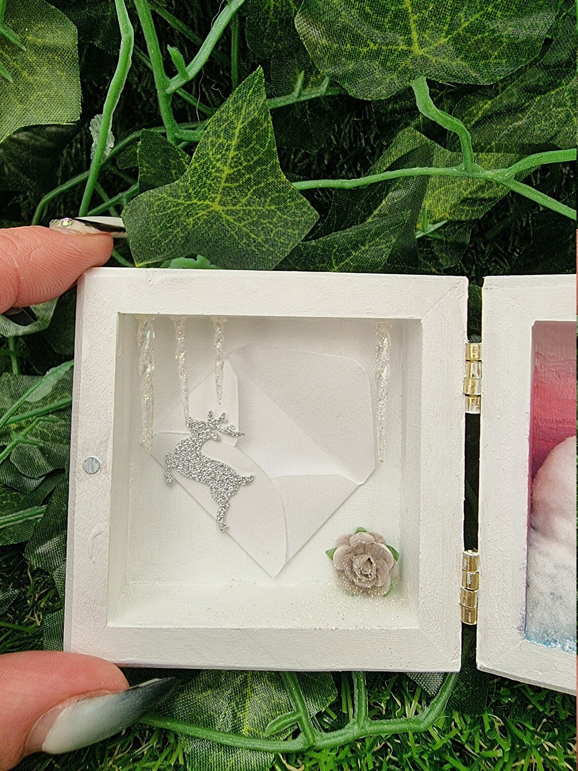 A small white square box with lid, open like a book. Detail is shown of the left side, which has icicles hanging down from the top. A miniature envelope with a reindeer motif in silver glitter is placed on the back wall. A grey rose is bottom right