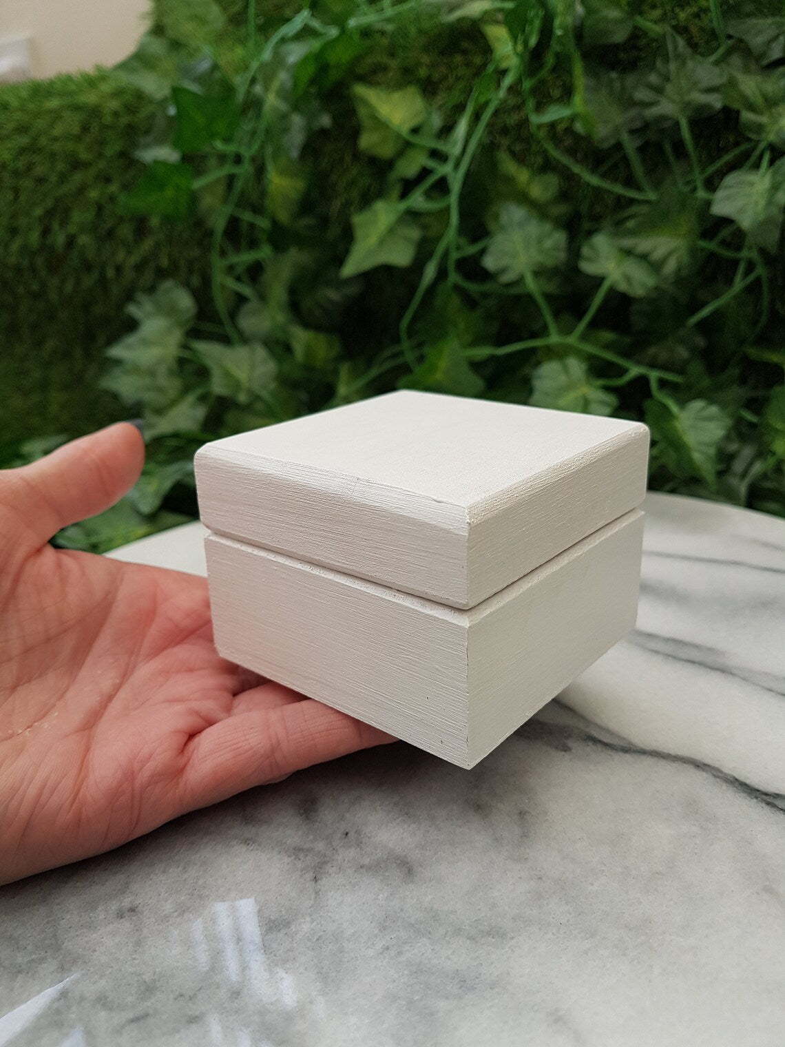 A small white wooden box is shown facing upwards in the open palm of a hand on a green background