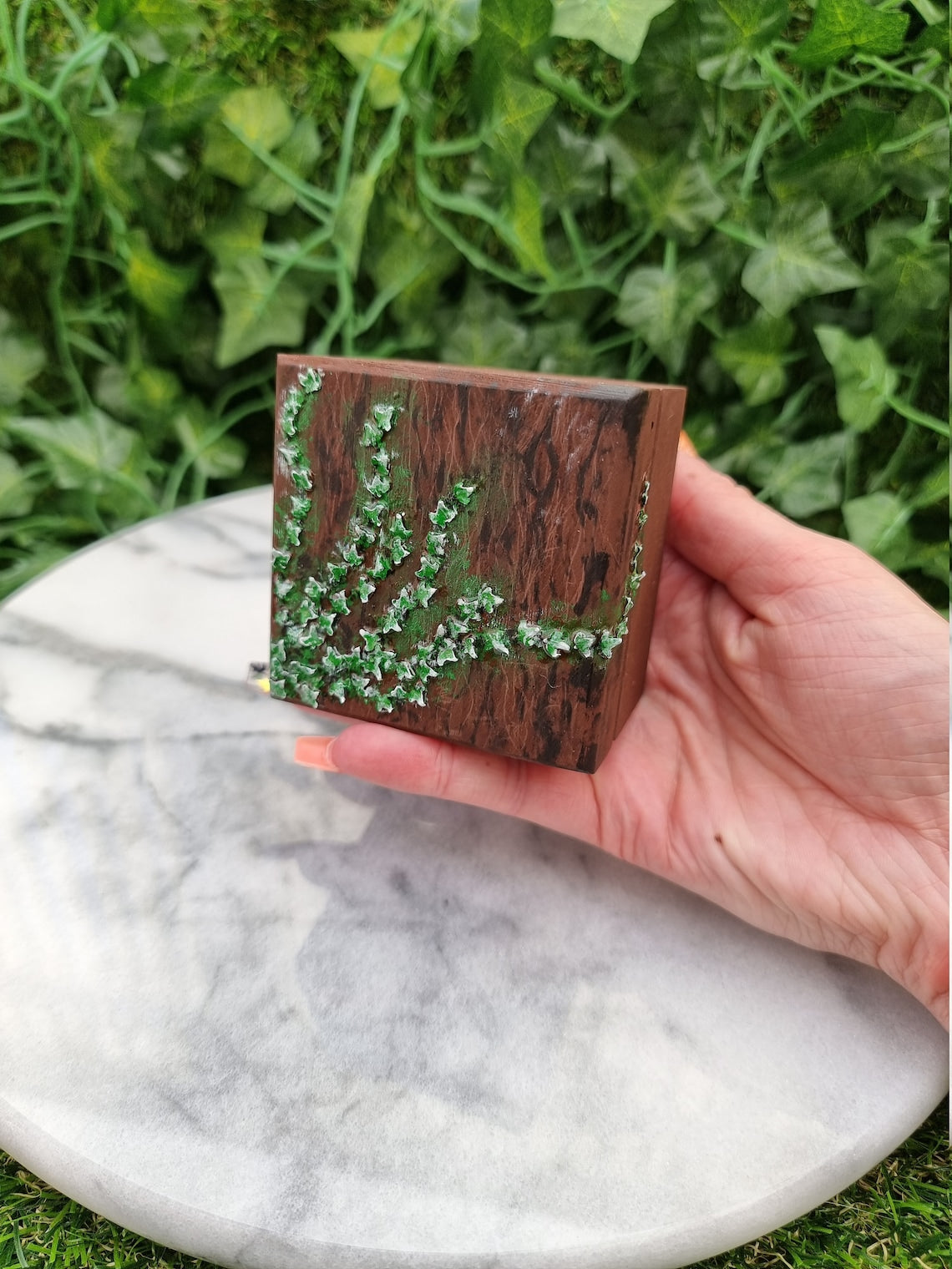 Small painted wooden box held in a hand against a marble background. The box has tiny green ivy leaves creeping across from left to right and around the side of the box