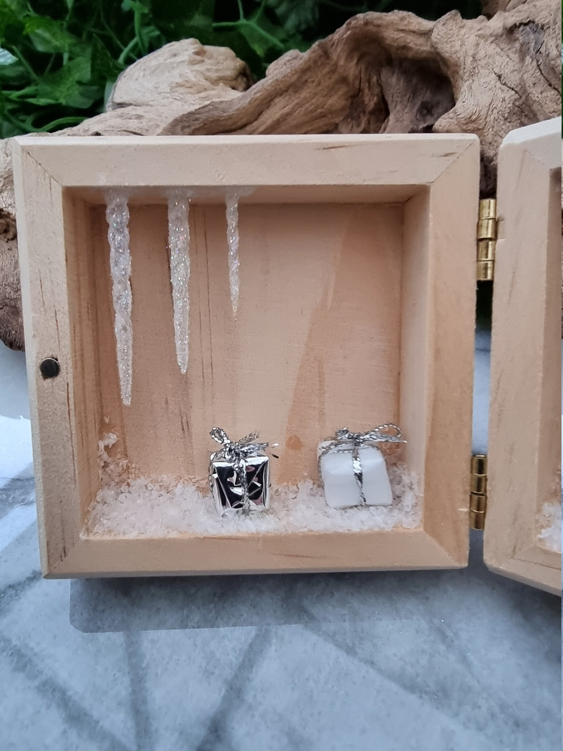 A small wooden box sits open like a book on a marble base. The box has snow, icicles and wrapped presents on the left hand side.