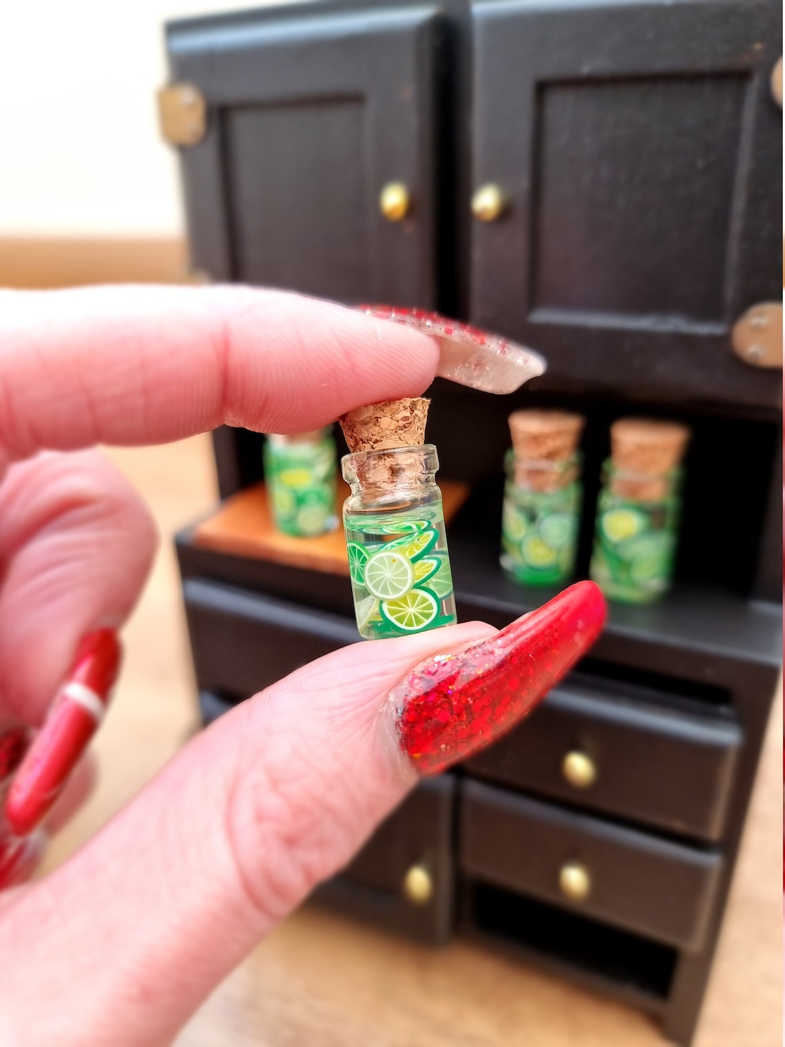 Single 1:12 scale miniature preserve jar with cork. Jar contains lime slices. Jar is held between thumb and finger. There is a black food cupboard in the background with more miniature jars of lime slices