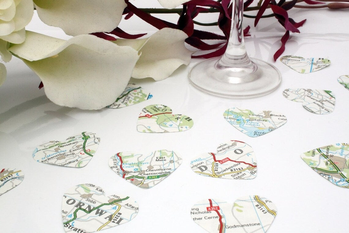 Map paper confetti pieces in heart shapes are scattered on a white background with a champagne flute, favour box and purple orchid bouquet in the background. The paper confetti is primarily white with green, blue and red colours