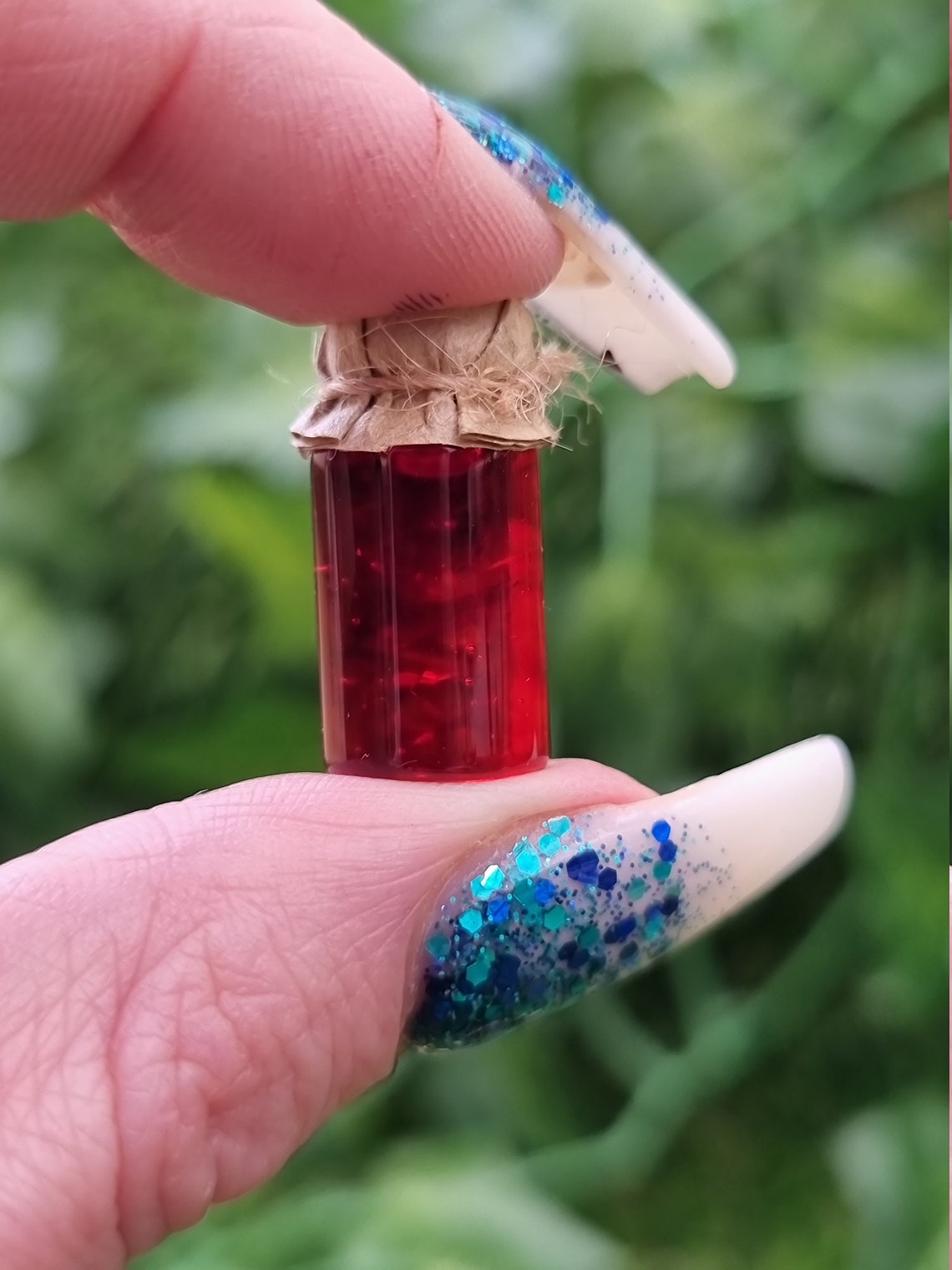 A miniature jar of crimson jam with brown paper lid is shown held between an index finger and thumb against a green background