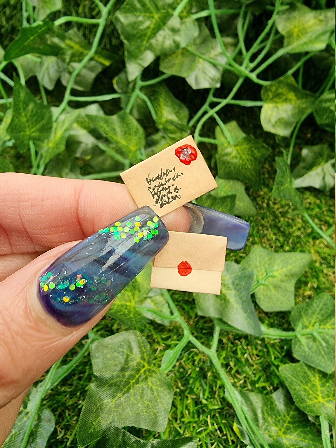 Two aged 1:12 scale miniature letters are held between thumbnail and finger. The letters are brown, both closed, one showing an address and stamp and the other showing the seal on the back. Green ivy is seen in the background