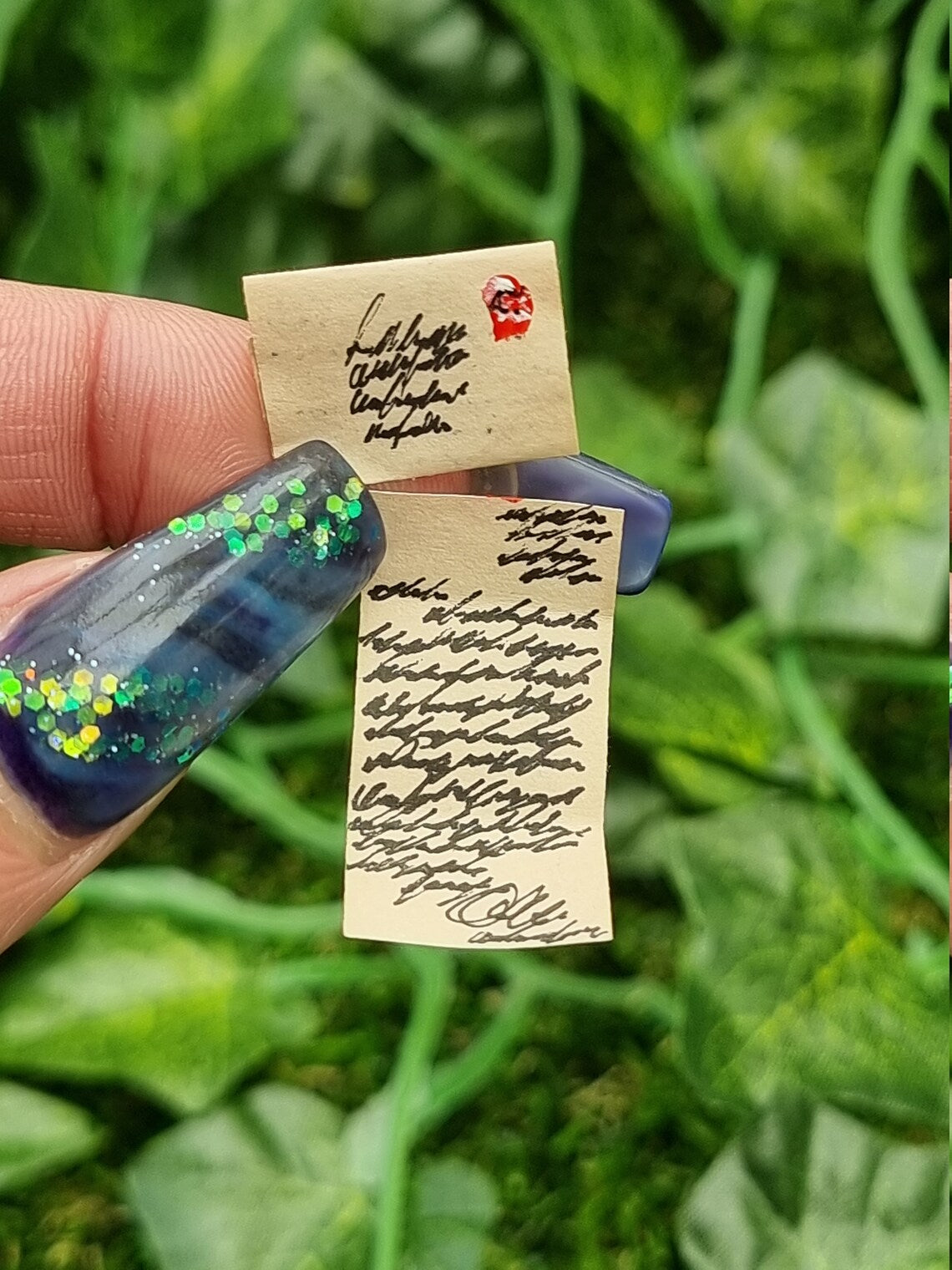 Two aged 1:12 scale miniature letters are held between thumbnail and finger. The letters are brown, showing black text. One letter has been opened, so has folds with letter text. The other shows a stamps and address.