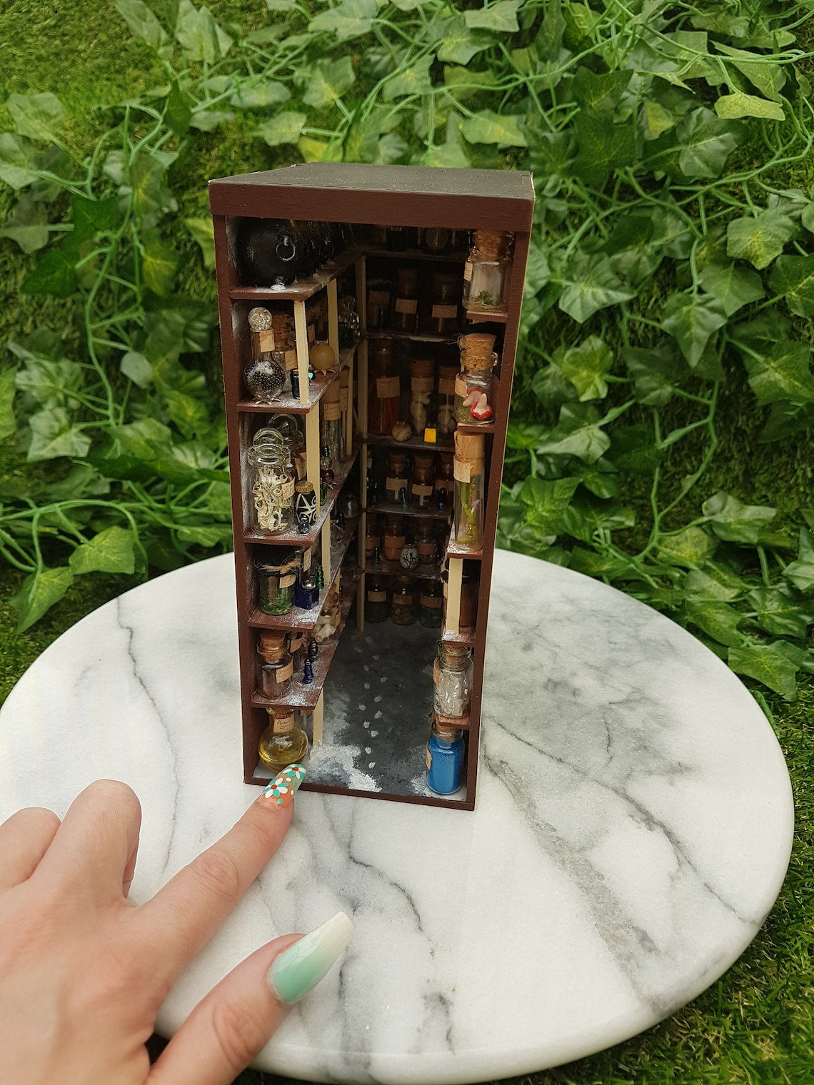 A small open fronted book nook shelf insert box sits on a marble slab. The box contains 5 sets of shelves up to the ceiling on all sides, each full of labelled glass vials and vessels. A finger points at a vial for scale