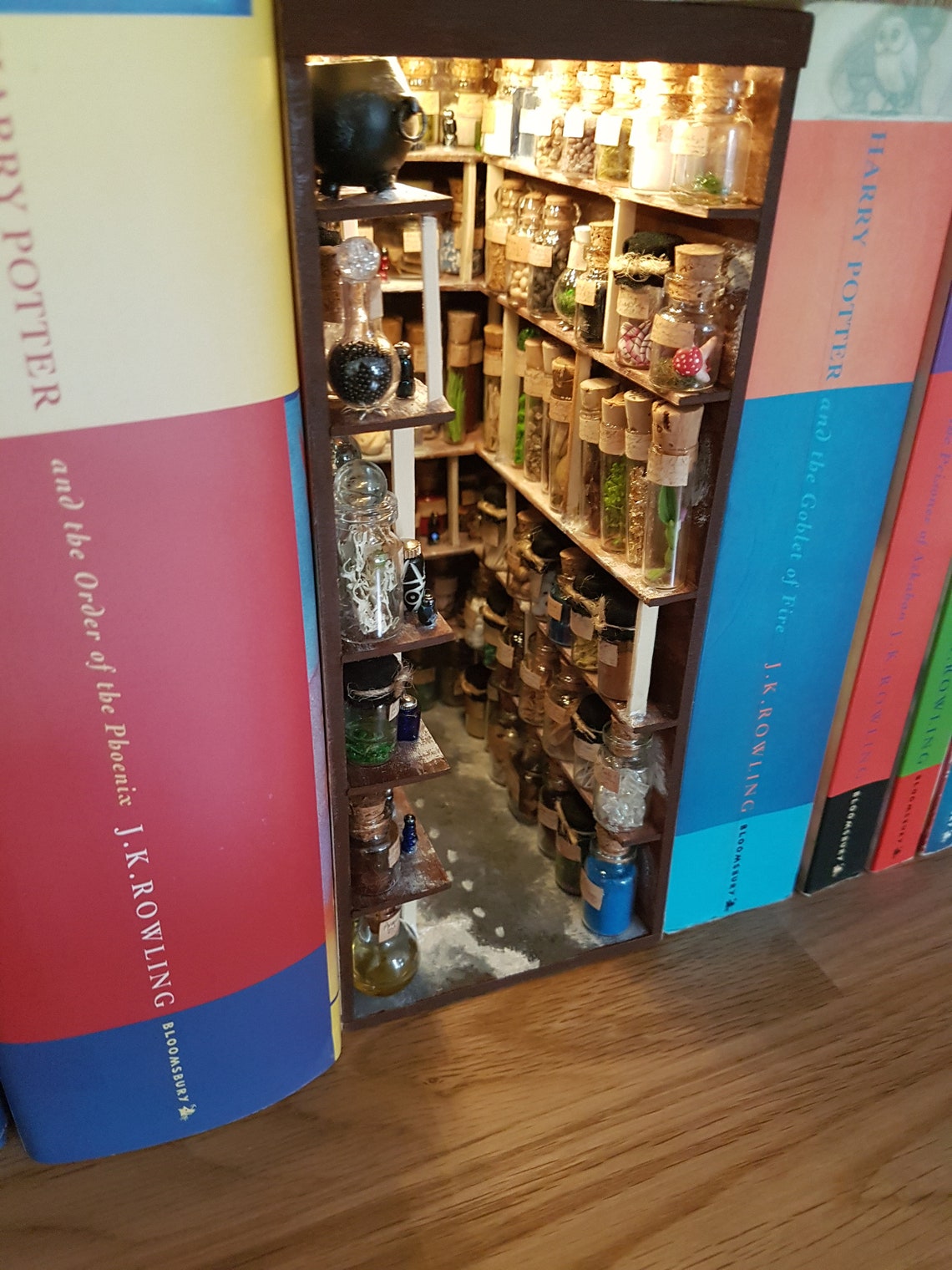 A small open fronted book nook shelf insert box sits between books of the same size on a bookshelf. The box contains 5 sets of shelves up to the ceiling on all sides, each full of glass vials and vessels. A row of cauldrons is seen in the top left.