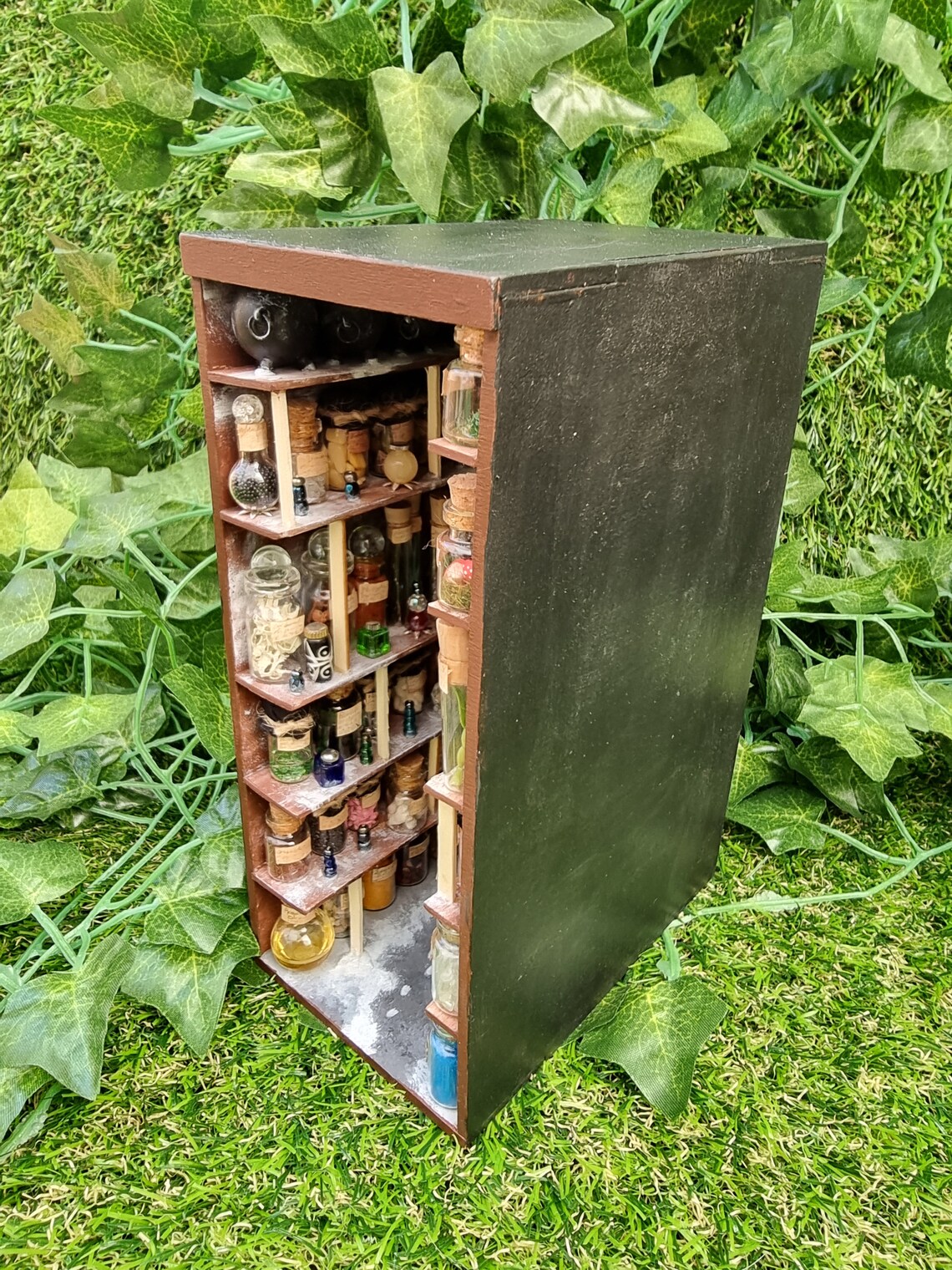 A small open fronted book nook shelf insert box sits on grass. The box contains 5 sets of shelves up to the ceiling on all sides, each full of labelled glass vials and vessels. A row of cauldrons can be seen ont he top shelf