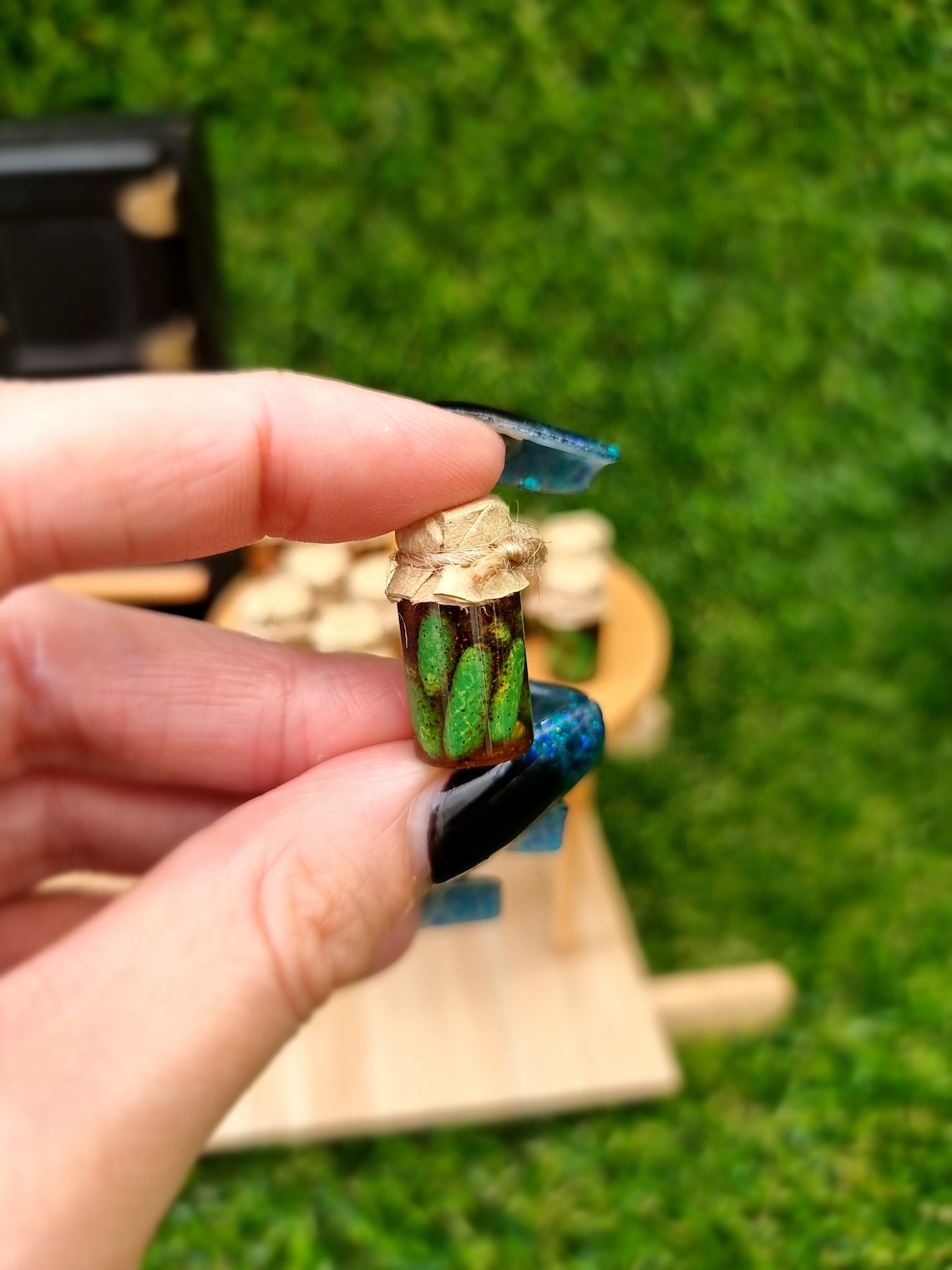 Single 1:12 scale miniature preserve jar with paper lid. Jar contains pickled gherkins. Jar is held between finger and thumb