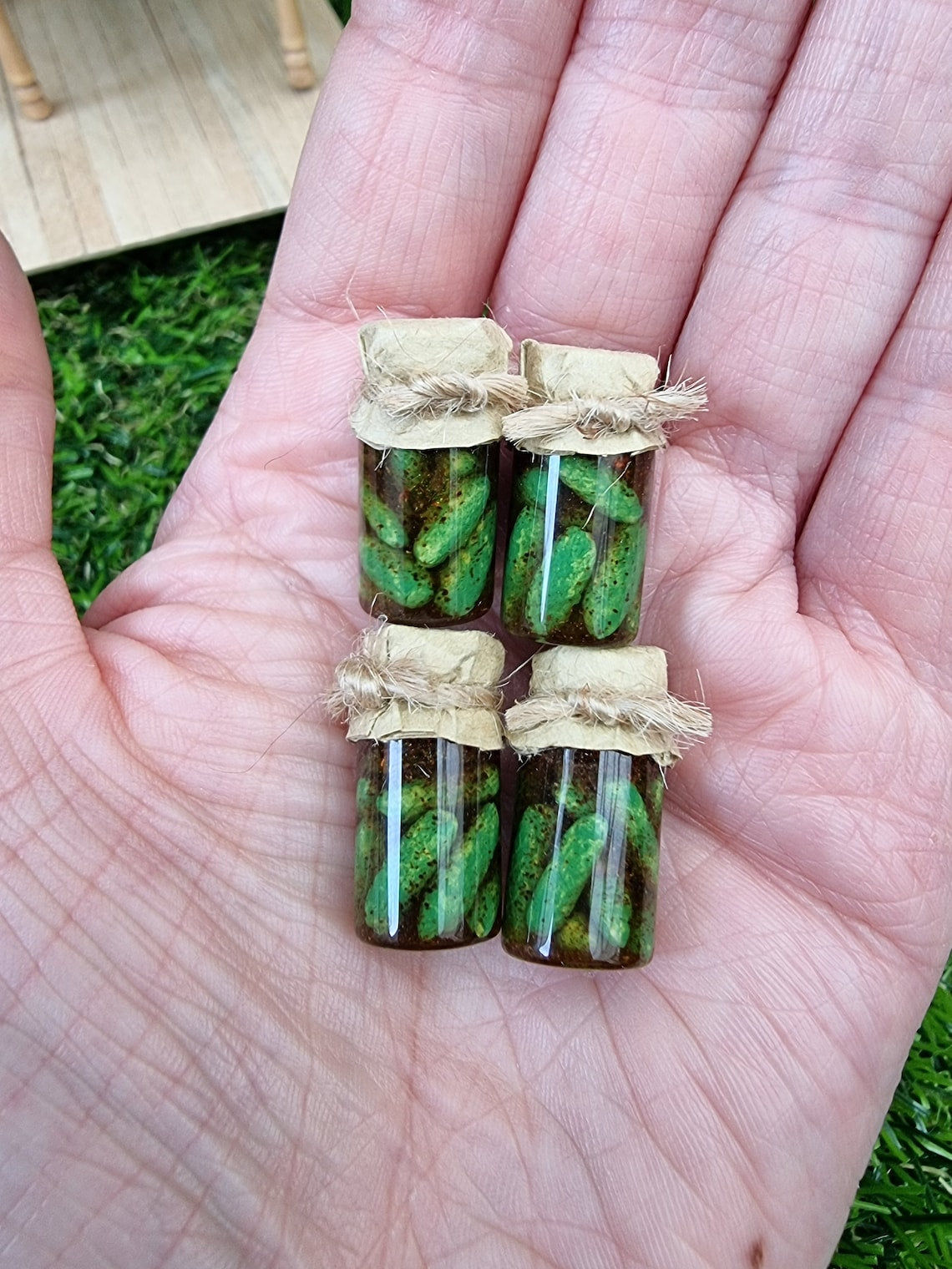 Several 1:12 scale miniature preserve jars with paper lids held in an open palm. Jars contain pickled gherkins