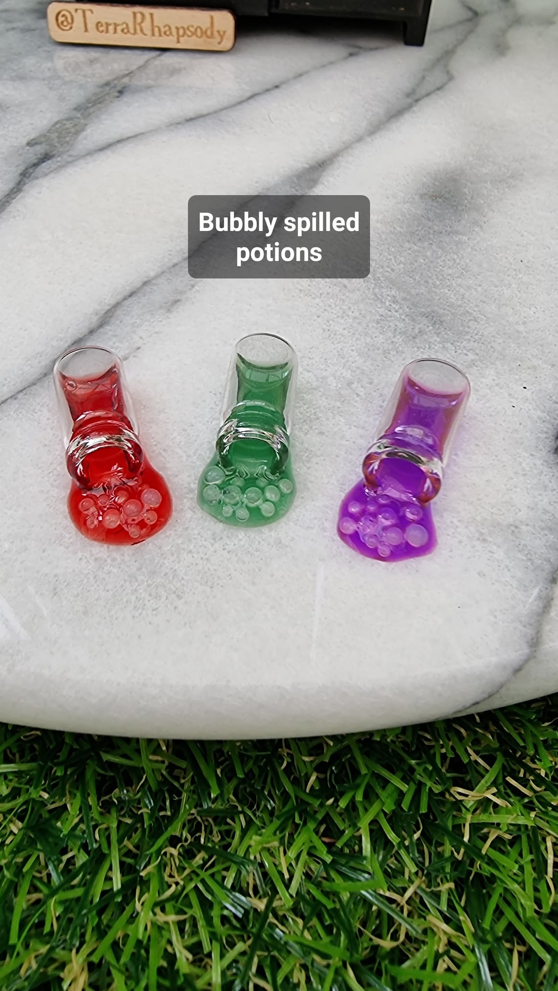 1:12 scale Miniature potion bottles are lying on their sides, appearing to have different coloured potions pouring our of them. The potion is bubbling. The bottles are lined up in front of a miniature dresser. Caption reads bubbly spilled potions
