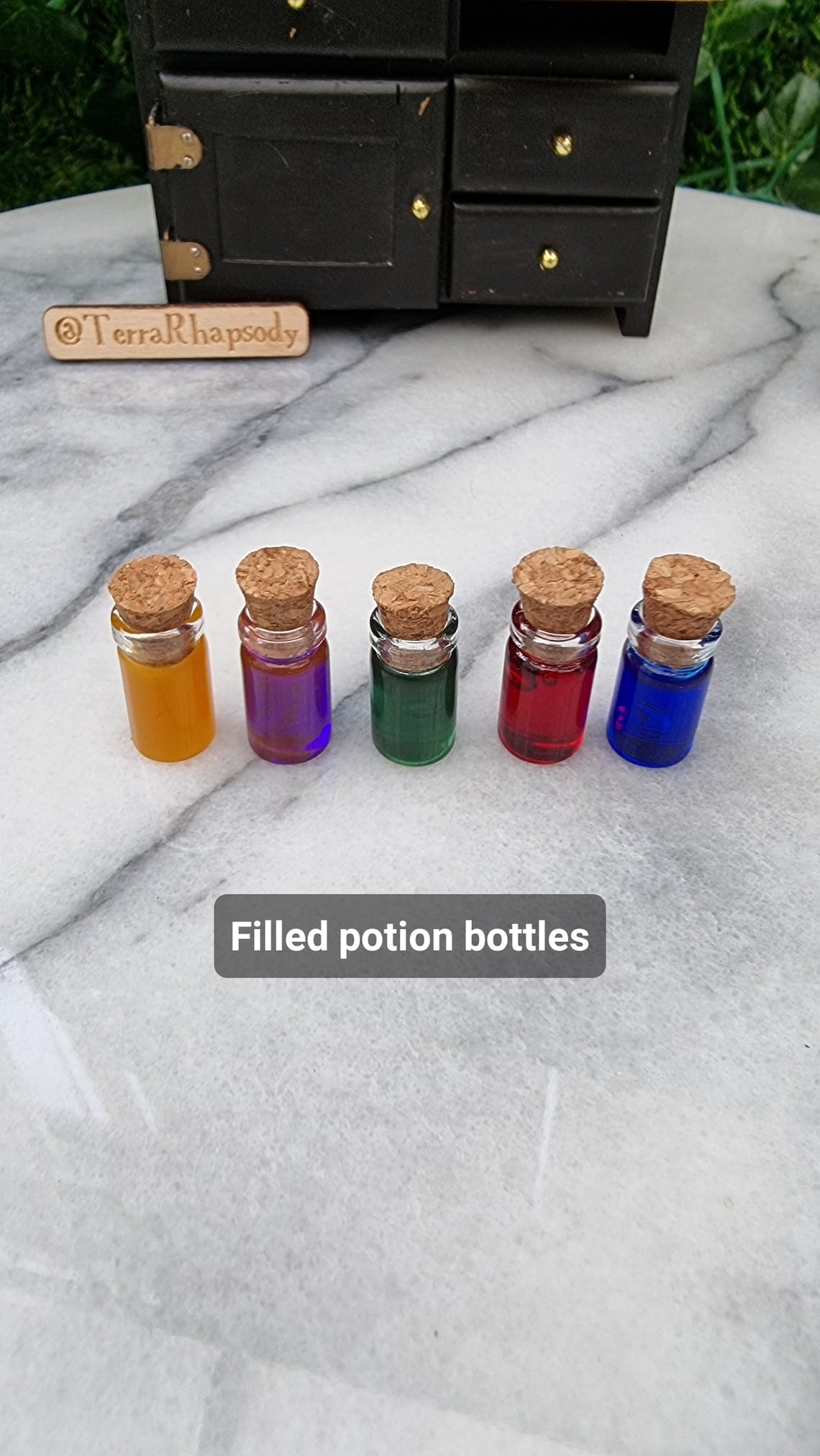 1:12 scale Miniature bottles with cork lids and different coloured resins are lined up in front of a black miniature dresser. The bottles are orange, purple, green, red and blue. A caption reads Filled potion bottles