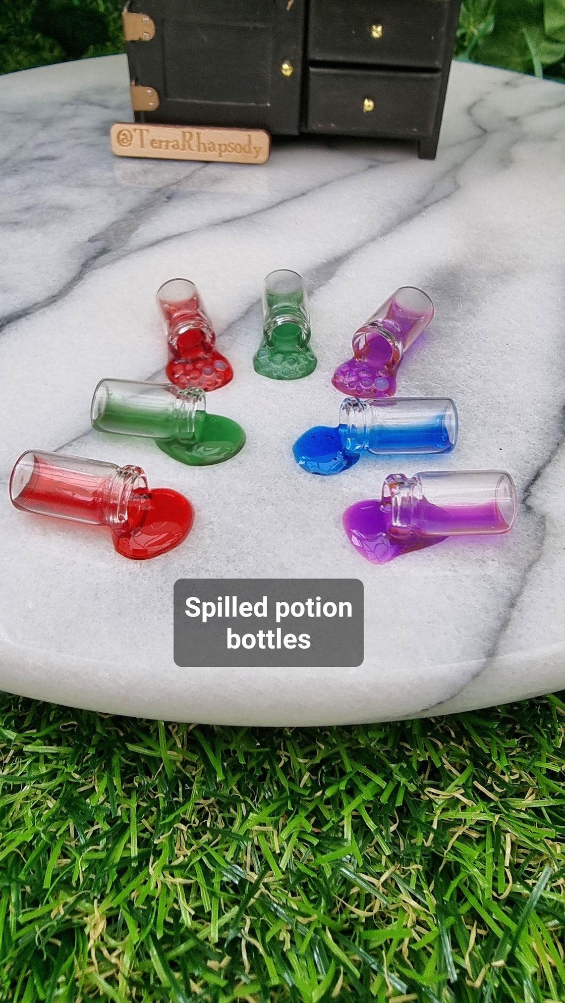 1:12 scale Miniature potion bottles with different coloured resins are lined up in front of a miniature dresser. The bottles are on their side, appearing to have coloured liquid pouring out of them