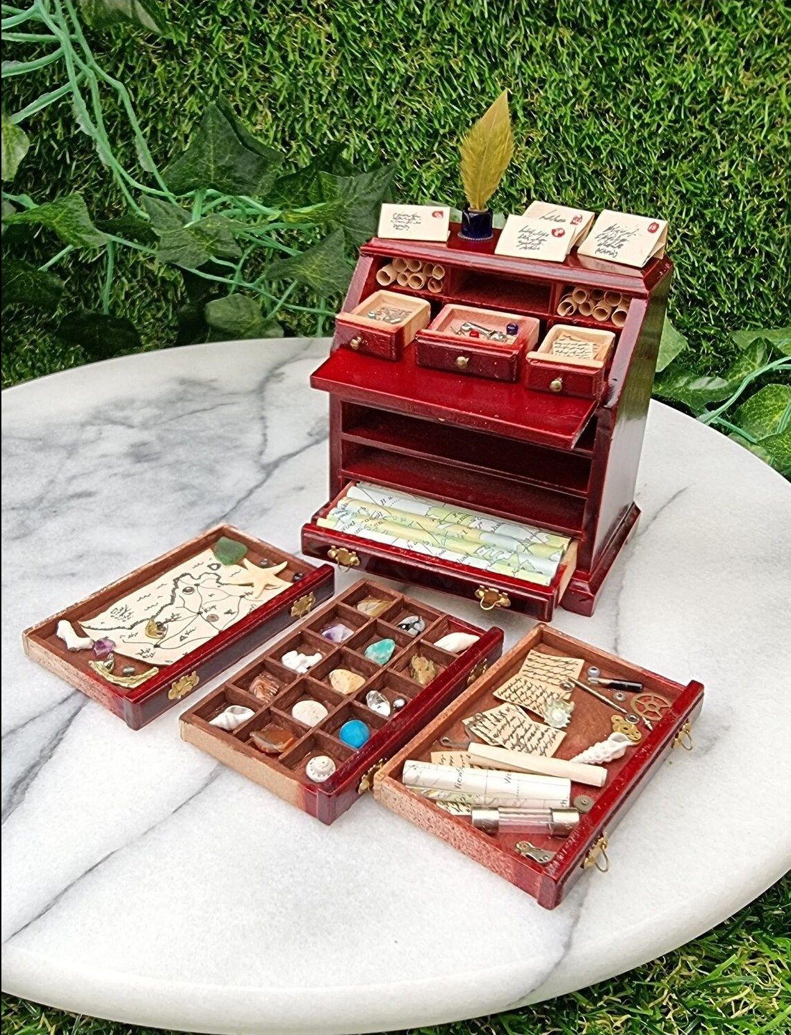 1:12 scale dollhouse brown desk has multiple drawers pulled out and sat in front of the desk. The filled drawers contain tiny maps scrolls, rocks, minerals and fossils, trinkets, letters and inkwell.