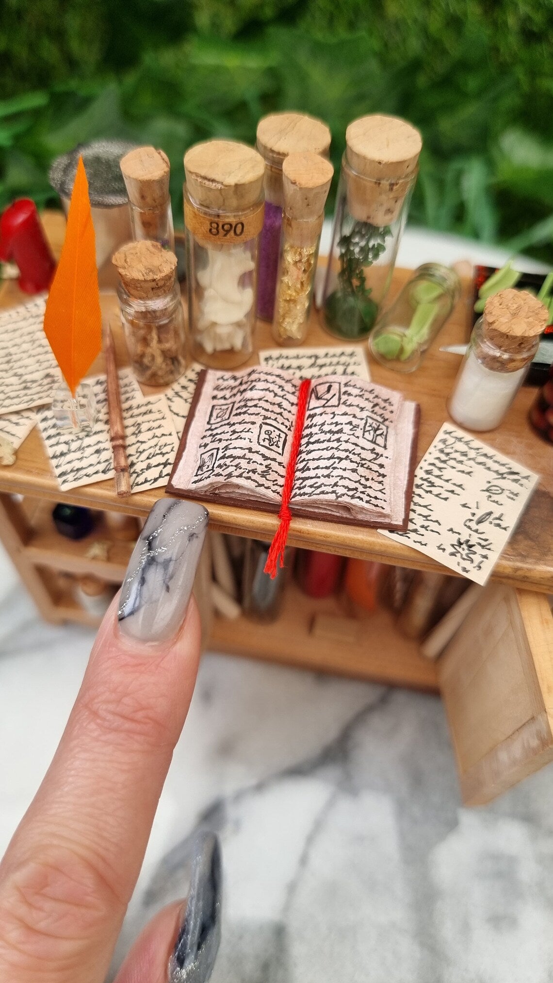 Top view of a 1:12 scale minature wooden sideboard. A finger points to an open book with a red bookmark, surrounded by an orange quill in an inkwell, wand, bottles of potion ingredients and handwritten parchments with tiny illustrations.