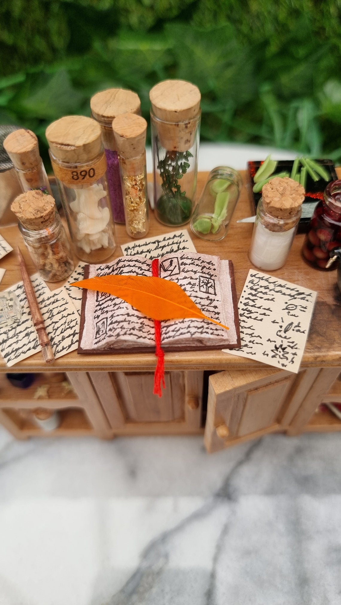 Top view of a 1:12 scale minature wooden sideboard, showing an open book with a red bookmark, orange quill, wand, bottles of potion ingredients and handwritten parchments with tiny illustrations. Leaves on a cutting board are seen on the other side