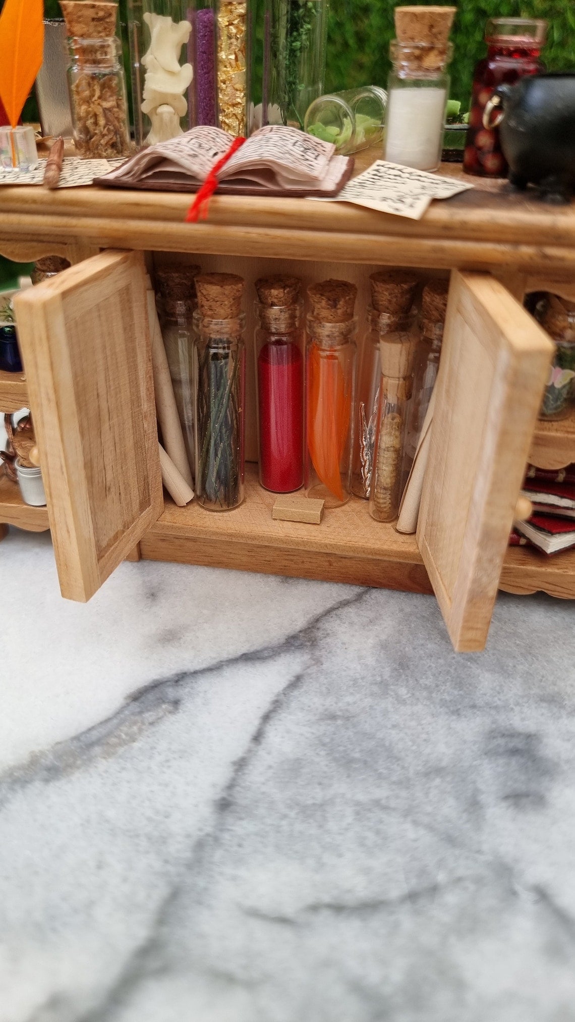 Back view of a 1:12 scale minature wooden sideboard with open cupboard doors revealing bottles of potion ingredients. The top has an open book with a red bookmark, orange quill, wand, cauldron and parchments