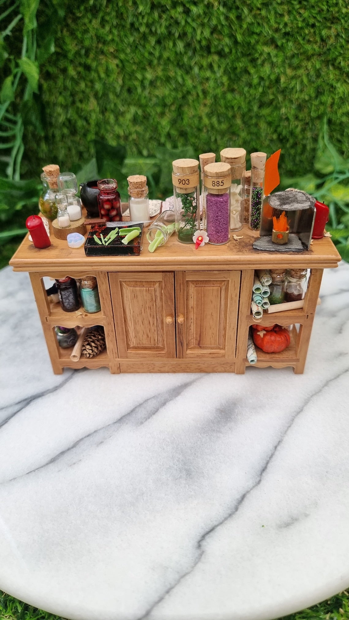 1:12 scale minature wooden sideboard with central cupboards and open shelves on the ends. The top is covered with parchment, wand, bottles of potion ingredients, open book, cauldron, notes, candles, sand timer, curios, inkwell and quill