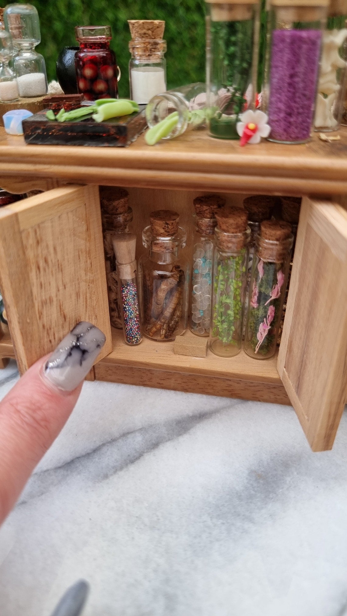 Front view of a 1:12 scale minature wooden sideboard. A finger points to a open cupboard filled with tall glass bottles of potion ingredients. The top hasgreen leaves on a cutting board, more filled bottle and sandtimers