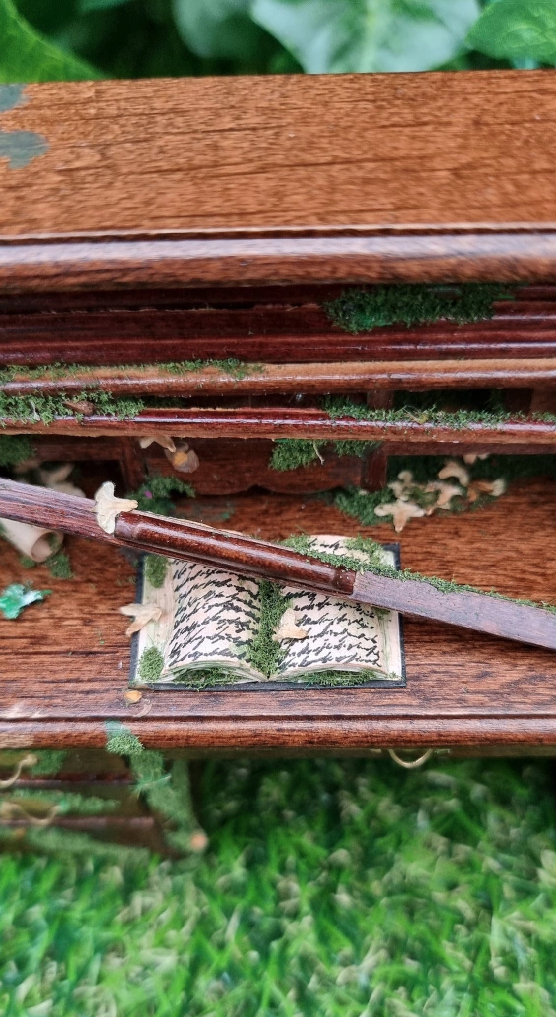 Desk top of a 1:12 scale miniature abandoned rolltop desk. The desk is brown with green moss and dead leaves strewn across it. An old book sits open, covered in moss, leaves and a broken wooden slat of the roll top