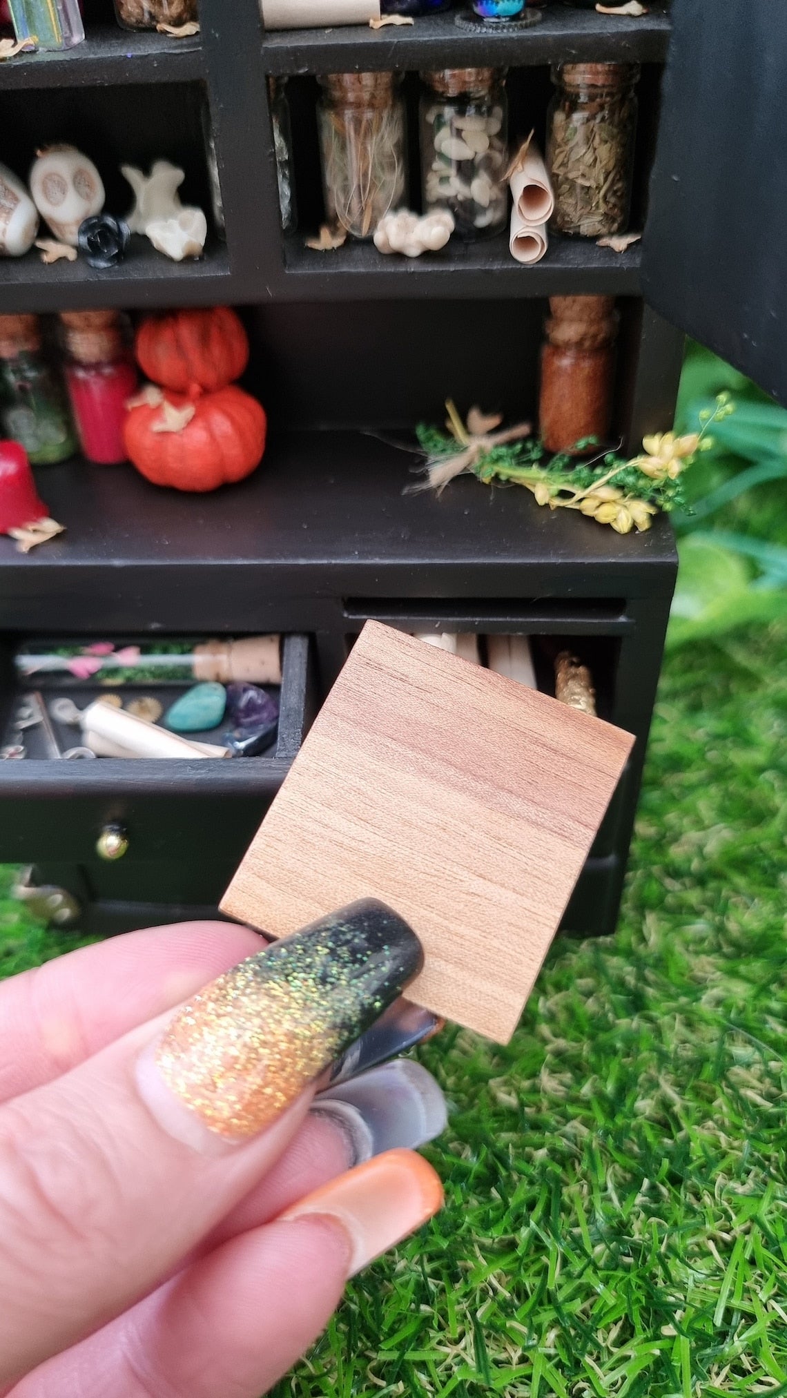 A hand holds a 1:12 scale wooden cutting board in front of a miniature black dresser. An open drawer contains flowers in a glass tube, key, parchments. The counter top has pumpkins and bottles. Open cupboards abovehave bottles, skulls and parchments