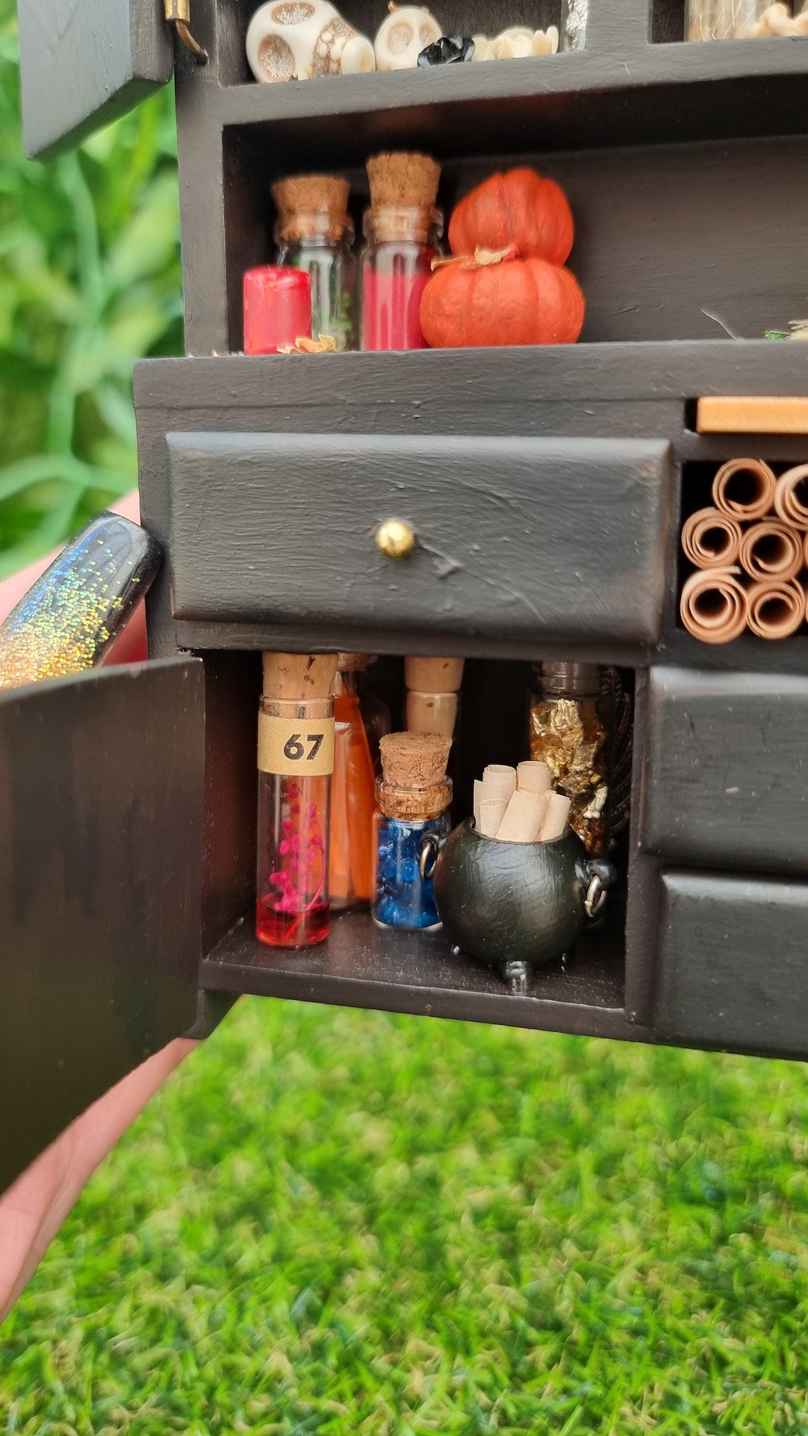 Hand holds 1:12 scale miniature black dresser with open cupboard door. Detail is shown of potion ingredients bottles and cauldron filled with rolls of parchments. The counter top has bottles, pumpkins and candle. Skulls are seen on a shelf above