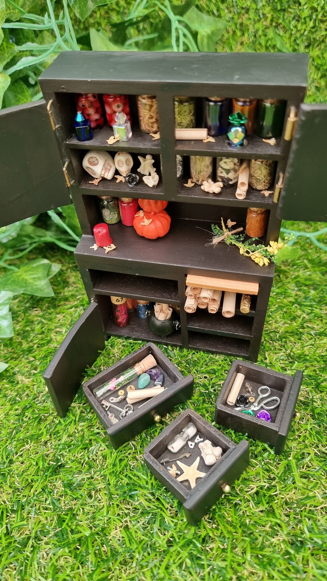 1:12 scale black dresser with drawers and cupboards. The cupboards contain bottles of potion ingredients, curios, bone and skulls. The counter top has pumpkins bottles, herbs and candle. The drawers contain parchment rolls, key and starfish