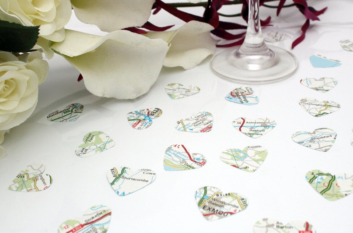 Map paper confetti pieces in heart shapes are scattered on a white background with a champagne flute, favour box and purple orchid bouquet in the background. The paper confetti is primarily white with green, blue and red colours