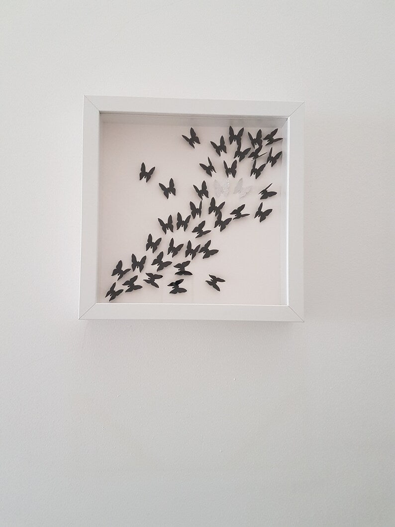Square white box frame showing a flurry of 3D black paper butterflies with two silver glitter butterflies, moving from the bottom left of the frame, through to the top right of the frame. Shown against a white wall background