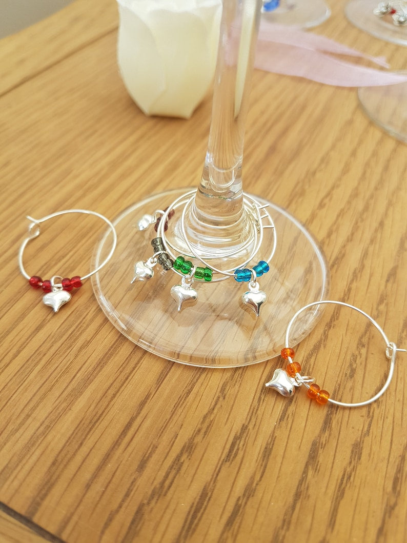 6 wine glass charms are arranged on a champagne flute base on a wooden background. The wine glass charms are round wire hoops, each with different coloured seed beads and a silver coloured puffy heart.