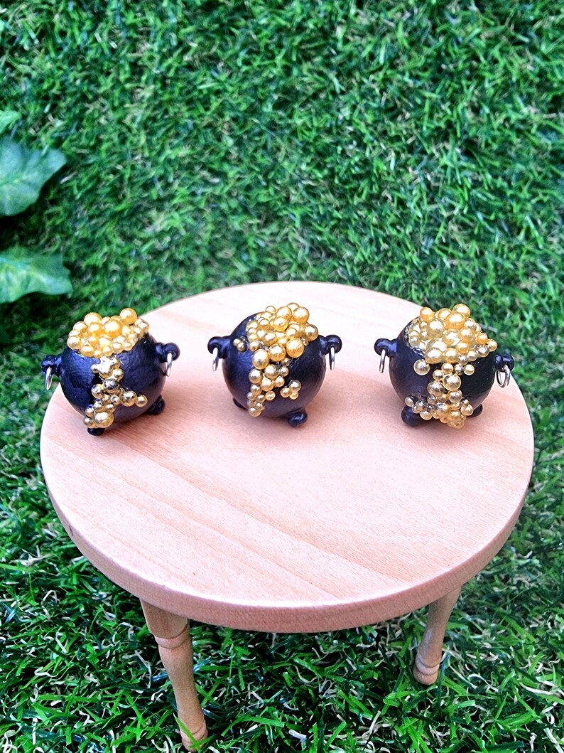 3 miniature black cauldrons overflowing with golden bubbles are displayed on a miniature wooden table against a green grass background