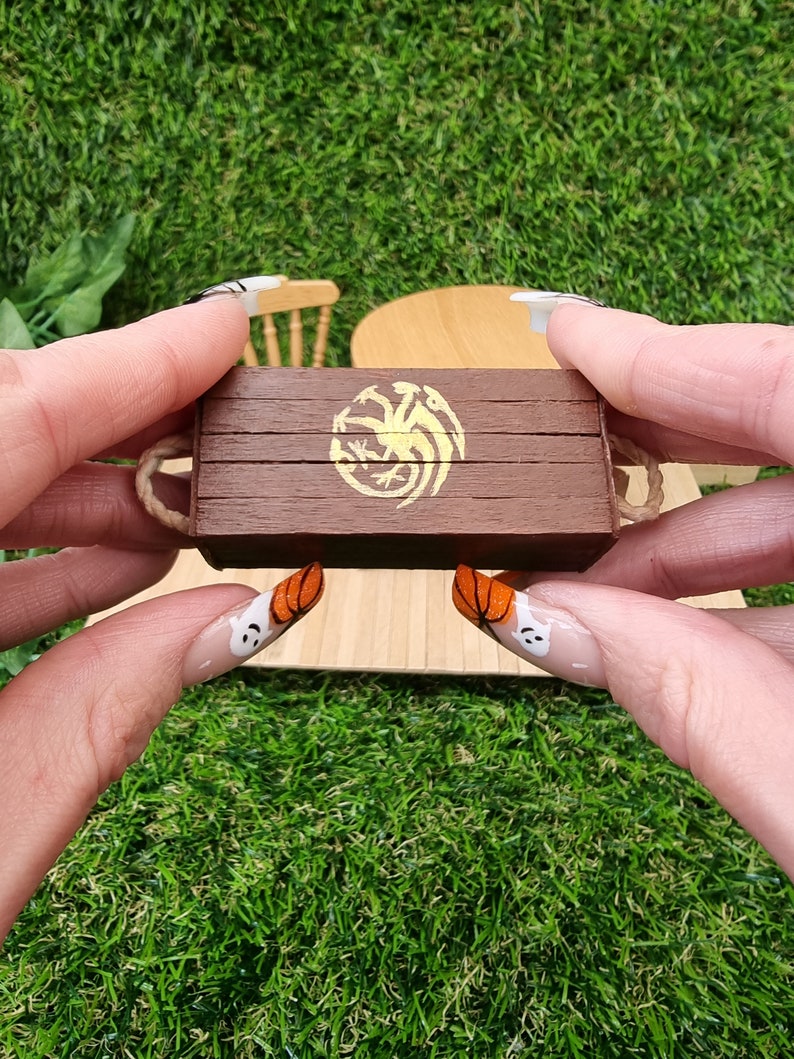 A hand holds a miniature brown lidded crate. The crate lid shows a gold Targaryen 3-headed dragon crest. The crate has rope handles on the short edge