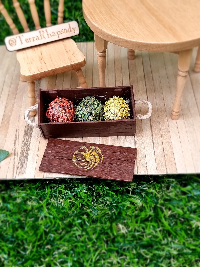 A miniature brown crate and lid contains 3 scaled dragon eggs- one red, one green and one yellow, all with a gold sheen. The crate lid shows a gold Targaryen 3-headed dragon crest. The crate has rope handles on the short edge