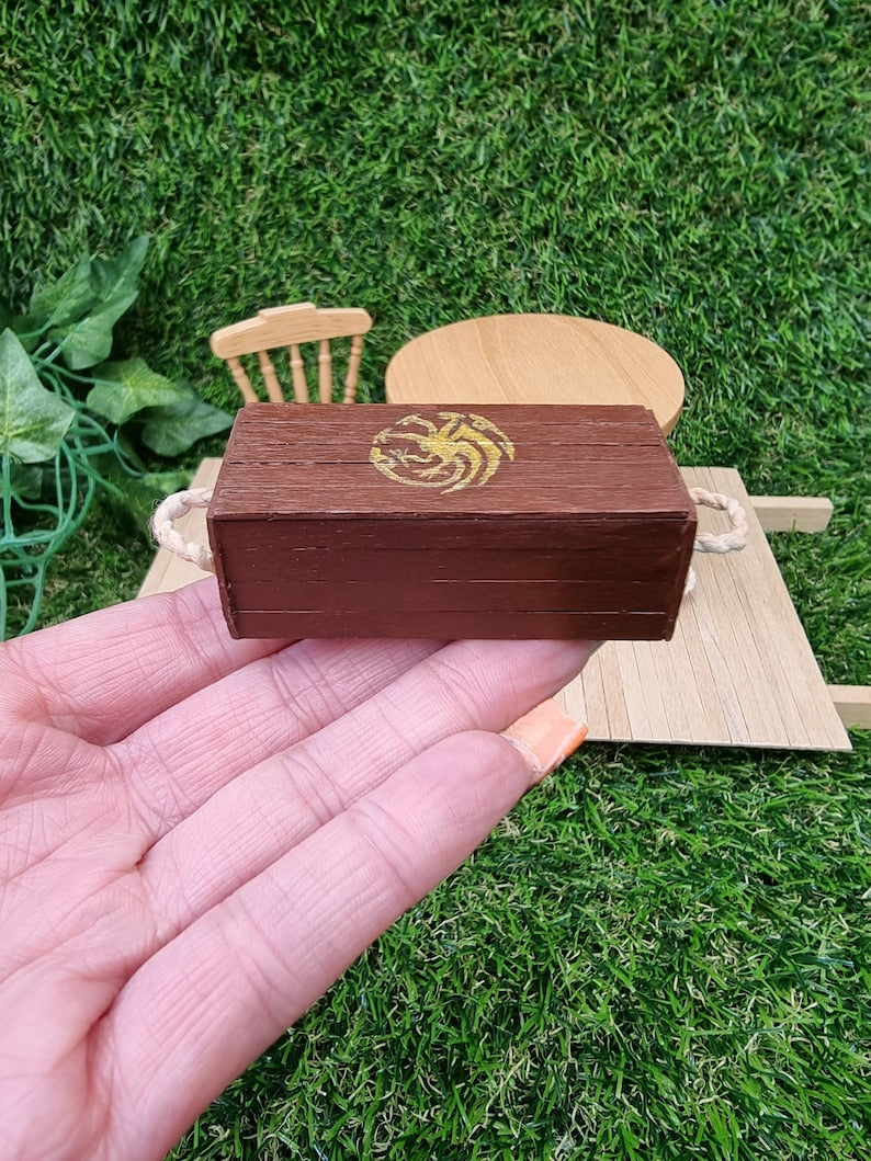 A hand holds a miniature brown lidded crate. The crate lid shows a gold Targaryen 3-headed dragon crest. The crate has rope handles on the short edge