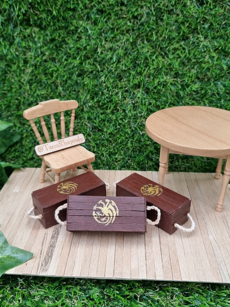 3 miniature brown lidded crates sit on a wooden base. The brown crates each bear a gold Targaryen 3-headed dragon crest. Displayed on a wooden base with other 1:12 scale hair and table.