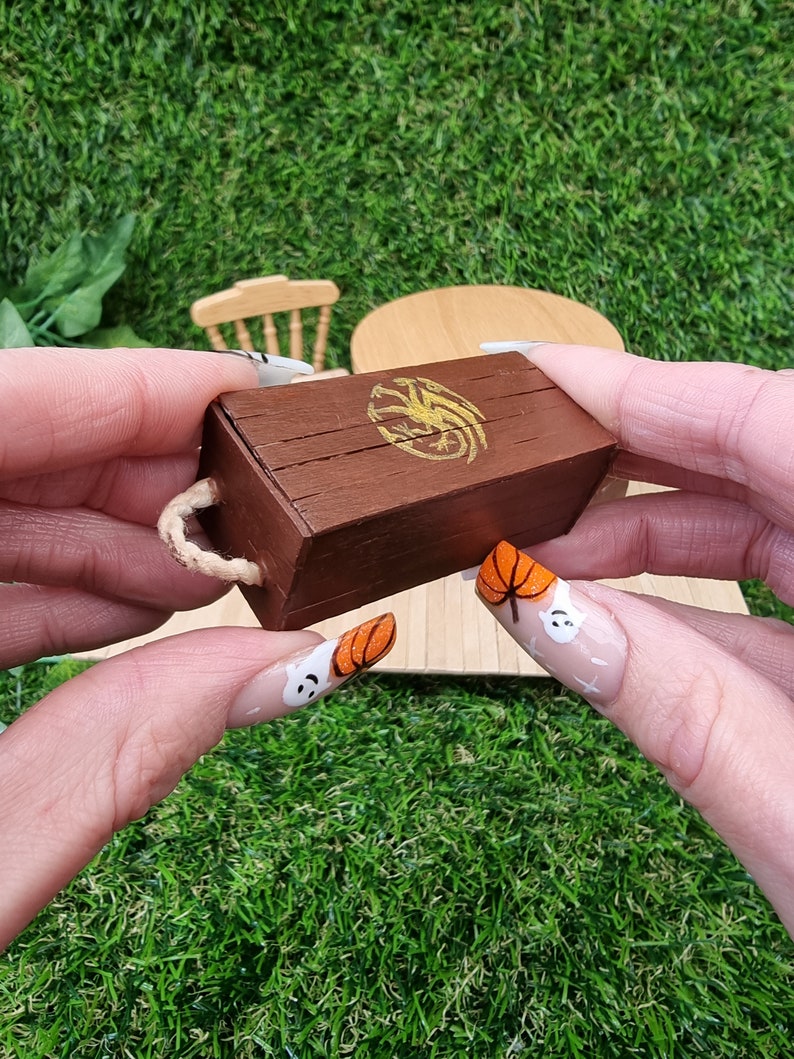 A hand holds a miniature brown lidded crate. The crate lid shows a gold Targaryen 3-headed dragon crest. The crate has rope handles on the short edge