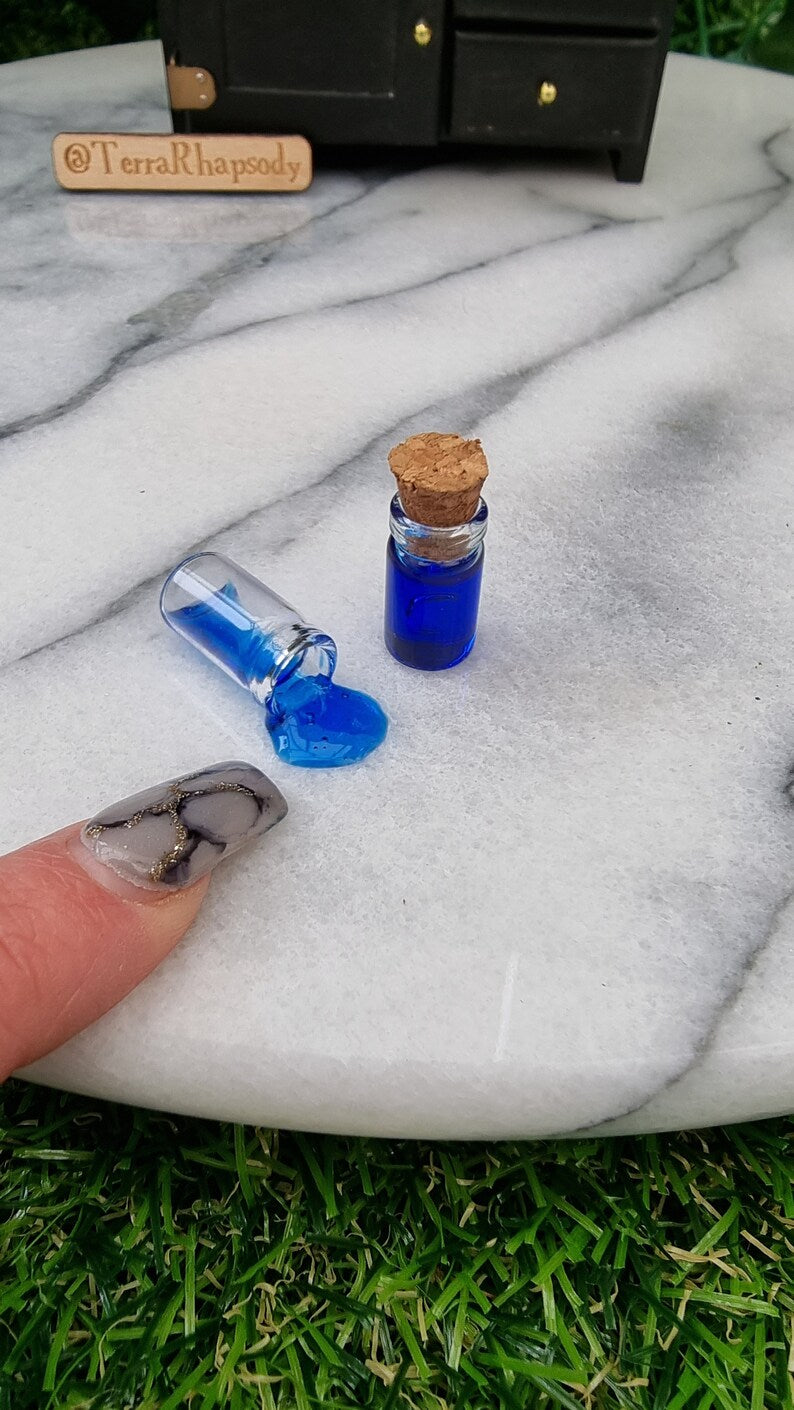 2 1:12 scale miniature bottles with blue potion are lined up in front of a miniature dresser. One bottle is filled and has a cork lid. 1 bottle is on its side, appearing to have blue potion pouring out of it. A finger points for scale