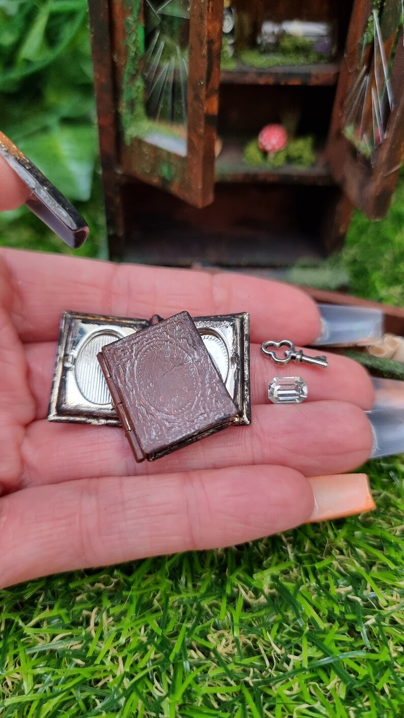 A hand holds a miniature brown book on top of an open book with a secret compartment, a key and a glass jewel. A miniature abandoned display cabinet sits in the background.