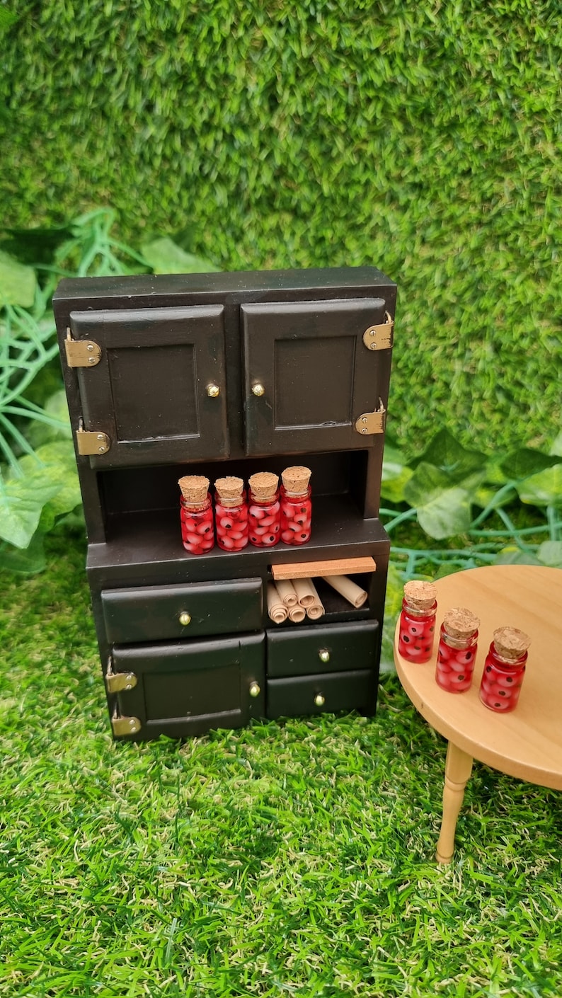 A 1:12 miniature black dresser and round wooden table holds 7 miniature glass jars containing eyeballs and red liquid. The miniature jars are topped with cork stoppers