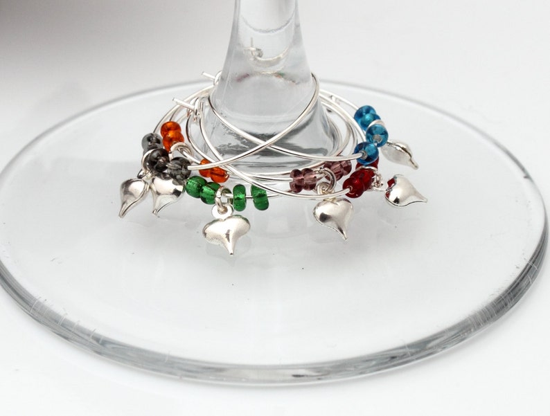 6 wine glass charms are attached to the base of a wine glass on a white background. The wine glass charms are round wire hoops, each with different coloured seed beads and a silver coloured puffy heart