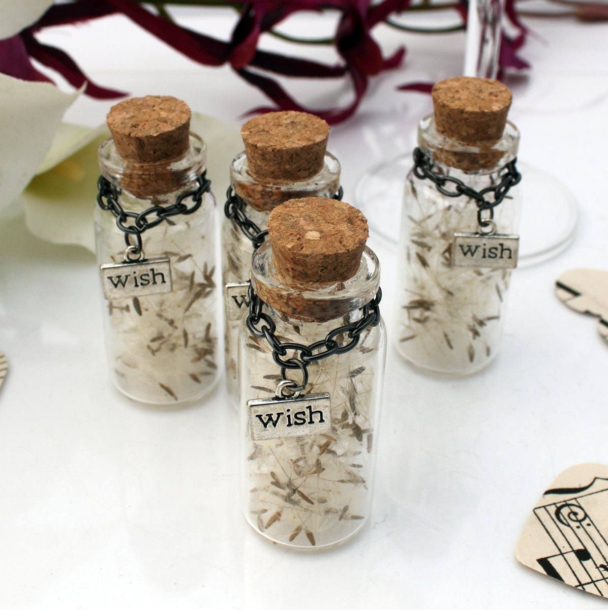 Four tall glass vials with cork lids containing dandelions seeds with silver coloured wish charms hanging from black chains around the jar necks. The jars are surrounded by paper confetti