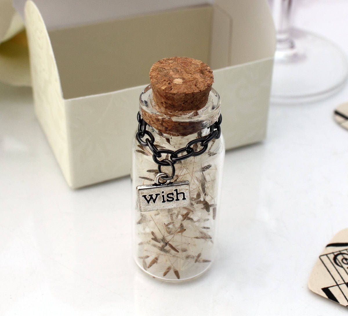 Single tall glass vial with cork lid containing dandelions seeds with silver coloured wish charms hanging from black chain around the jar neck. There is a favour box in the background