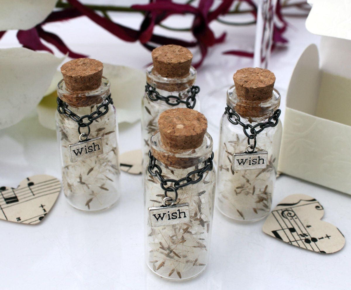 Four tall glass vials with cork lids containing dandelions seeds with silver coloured wish charms hanging from black chains around the jar necks. The jars are surrounded by paper confetti and wedding favour box