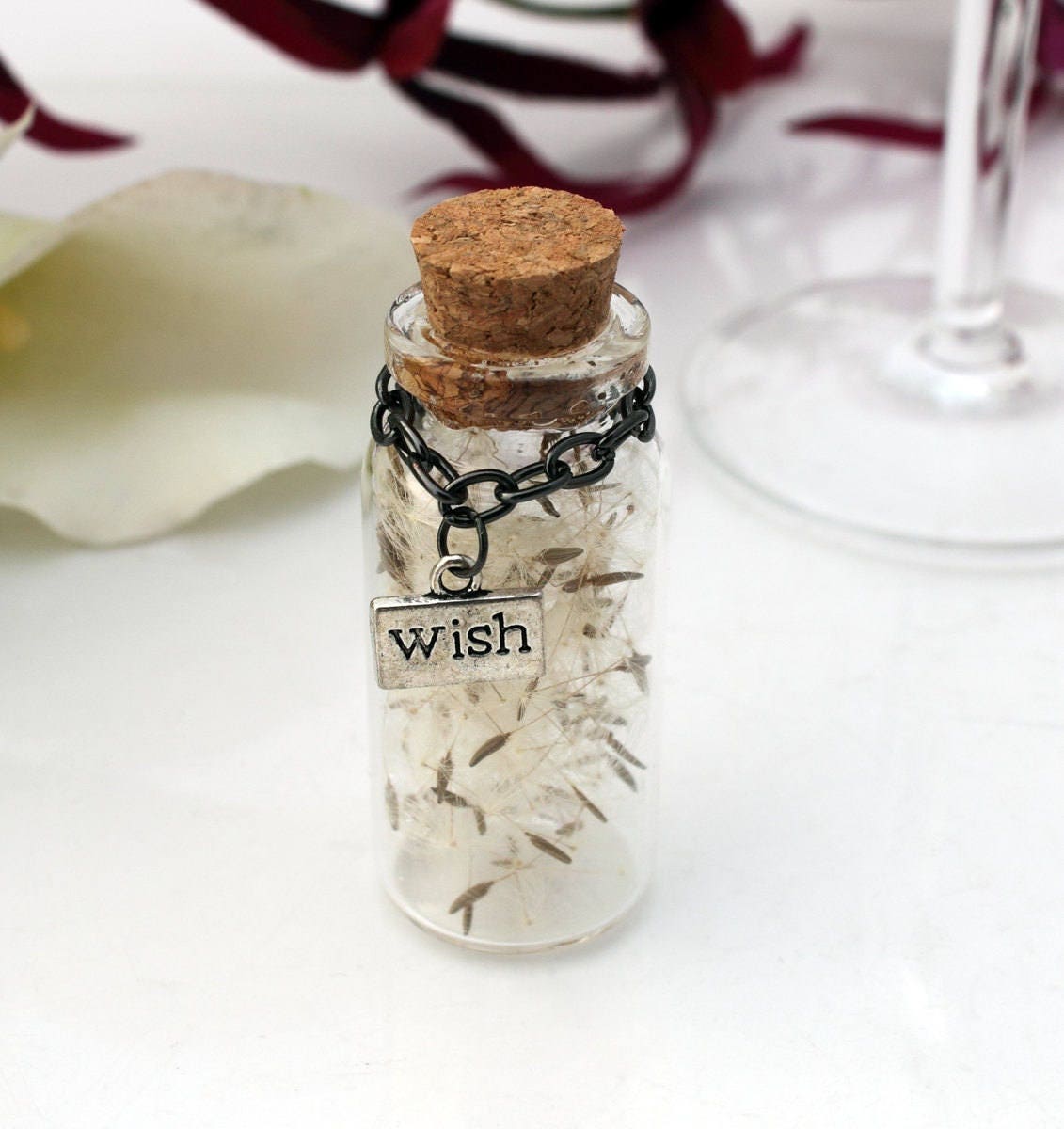 Single tall glass vial with cork lid containing dandelions seeds with silver coloured wish charms hanging from black chain around the jar neck. There is a wedding favour box in the background